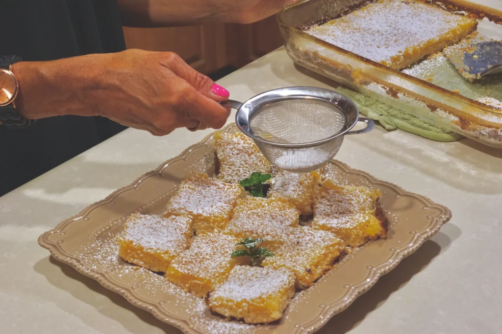 Lemon cream cheese bars being dusted with powdered sugar on a decorative plate