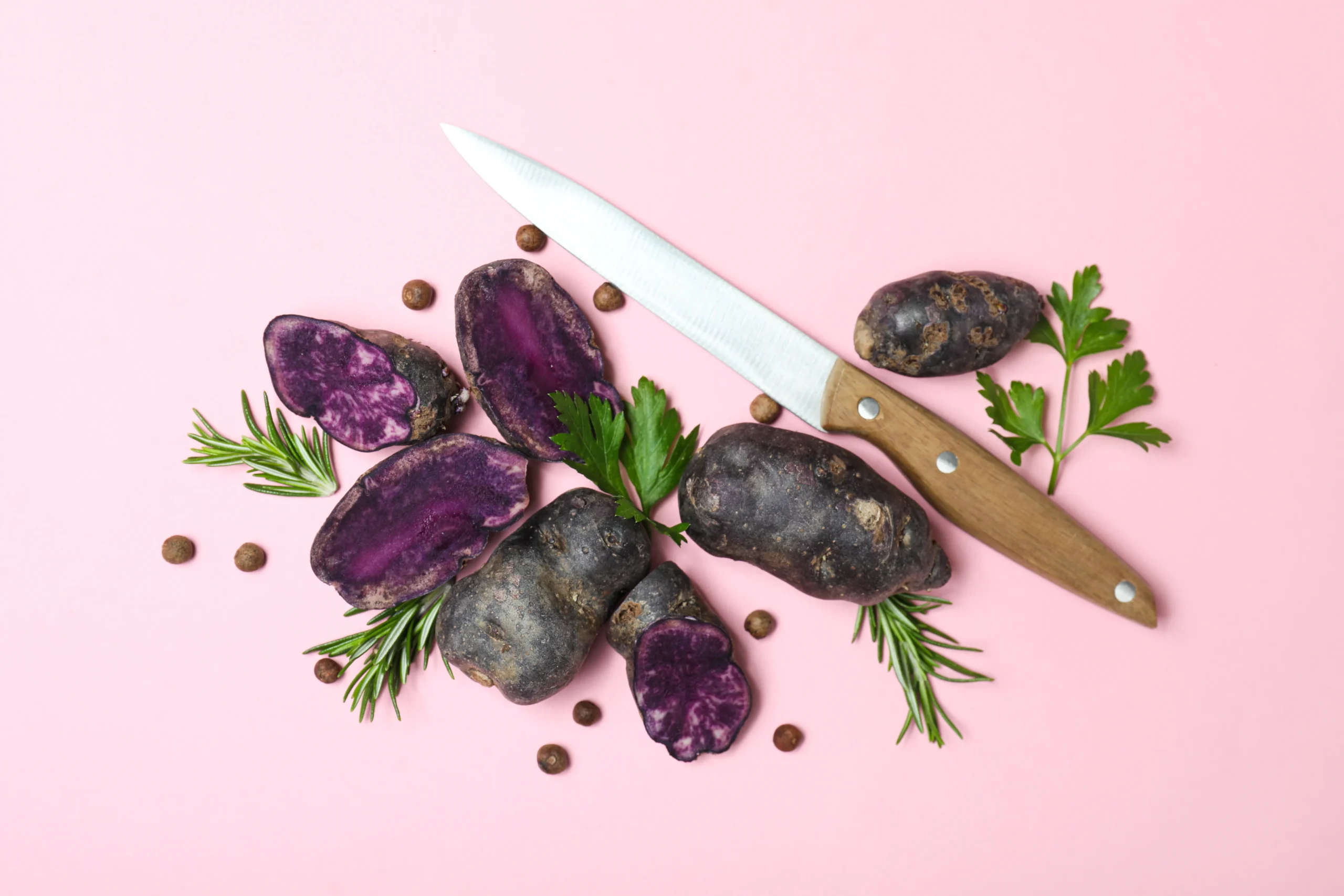 Purple sweet potatoes with a kitchen knife and fresh herbs on a pink background