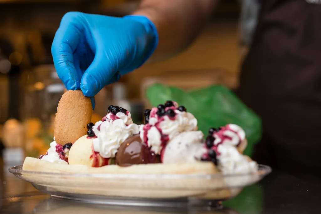 A close-up of a gloved hand placing a biscuit onto a banana split topped with whipped cream and berry sauce